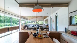 A modern dining area with a wooden table, chairs, and a fruit bowl. There is a kitchen area with a red toaster and a wine bottle. The space has large windows and a ceiling with a hanging light fixture. - VILLA WITH LAND IN LLANES (La Pereda)