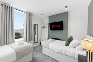 Ein modernes Hotelzimmer mit zwei Betten, einem großen Fenster mit Vorhängen, einem am Wand montierten Flachbildfernseher und einem Nachttisch mit Lampe.