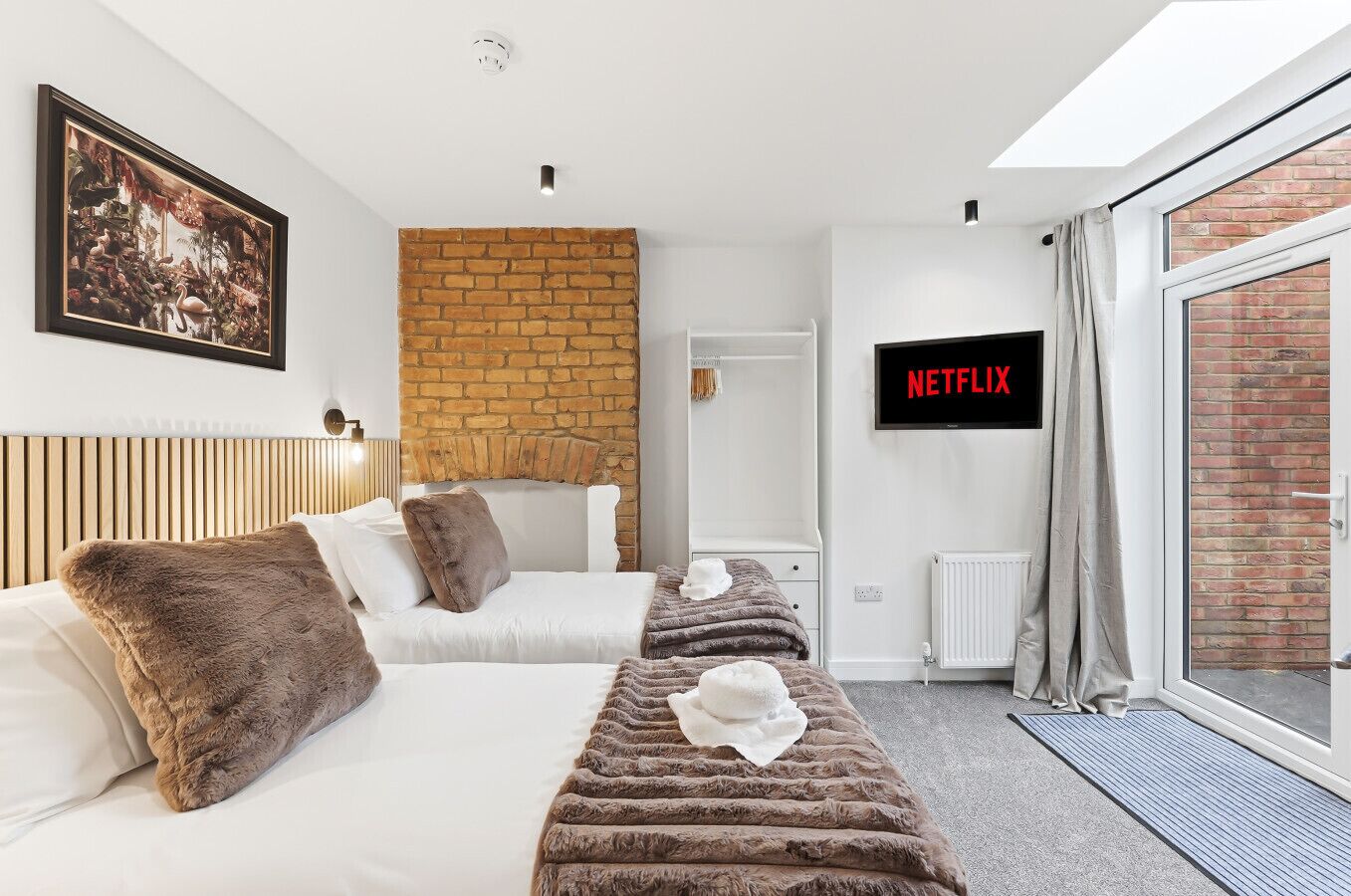 A modern hotel room with a brick accent wall, a large window with curtains, a bed with fluffy pillows, a bedside table, and a wall-mounted TV displaying 'NETFLIX'.