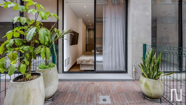 A modern building entrance with large potted plants, a visible bedroom with a bed and curtains, and a metal fence. - NOK Stylish 1-bedroom With Terrace on Calle 94 (Bogotá)