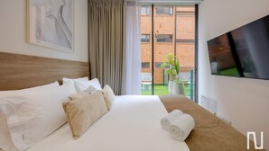 A hotel room with a large bed, a flat-screen TV, a view of a brick building, and a balcony with a potted plant. - NOK Stylish 1-bedroom With Terrace on Calle 94 (Bogotá)