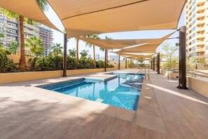 A swimming pool area with a tiled deck, palm trees, and a building in the background. - Stayshort - 1BR in JVC That Sleeps 4 With Amazing Amenities (Dubai)