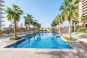A swimming pool with palm trees and lounge chairs, surrounded by buildings. - Stayshort - 1BR in JVC That Sleeps 4 With Amazing Amenities (Dubai)