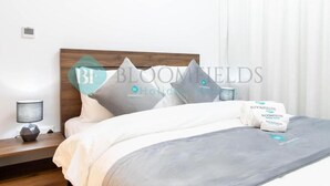 Una cama bien hecha con almohadas y una colcha, una mesita de noche con una lámpara, y una ventana con cortinas.