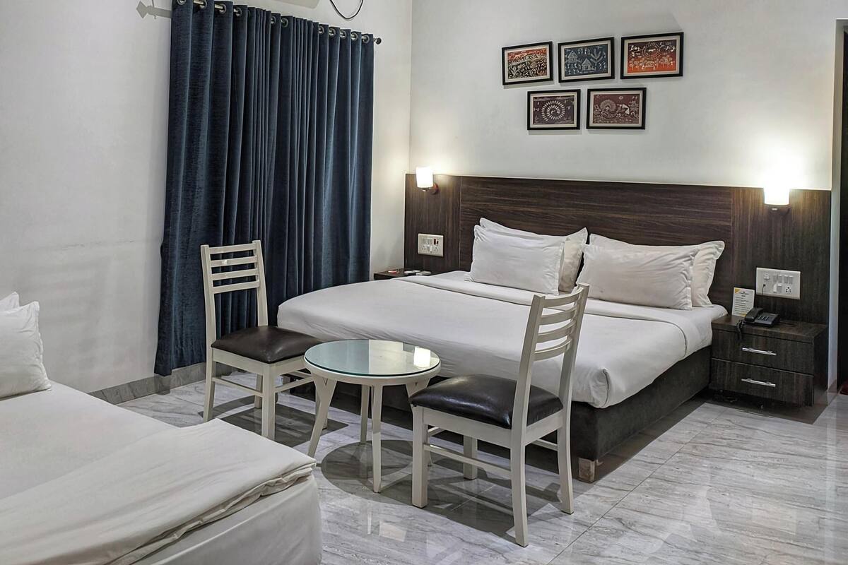 Habitación de hotel con dos camas, una mesita redonda pequeña, una silla, una mesita de noche y una pared con cuadros.