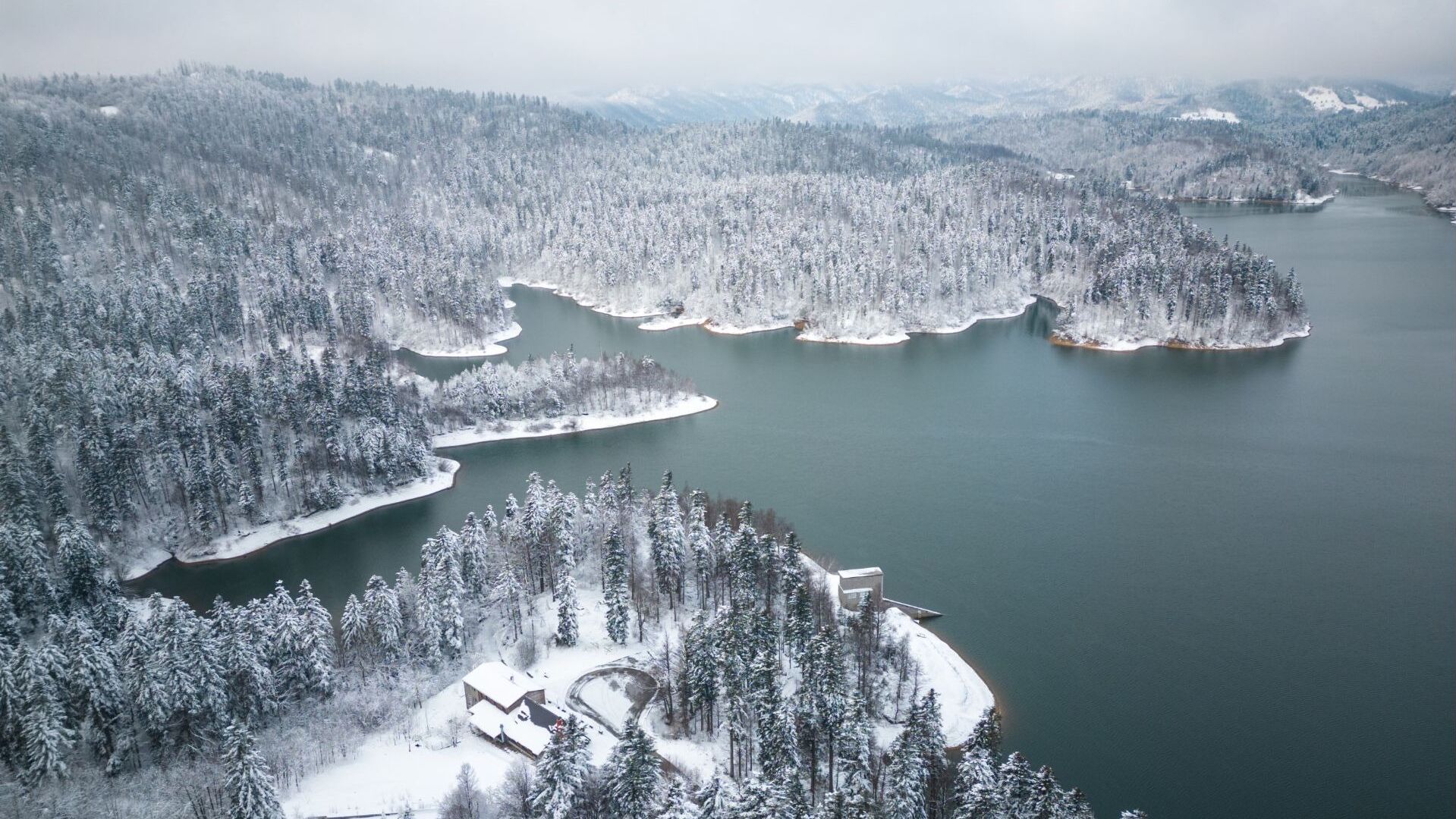 An aerial view of a snow-covered forest with a lake and a small building with a snow-covered roof.