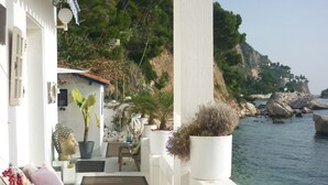 Un sentiero stretto conduce a un balcone con vista su una zona costiera caratterizzata da pareti bianche, piante in vaso e uno sguardo sul mare e le colline.