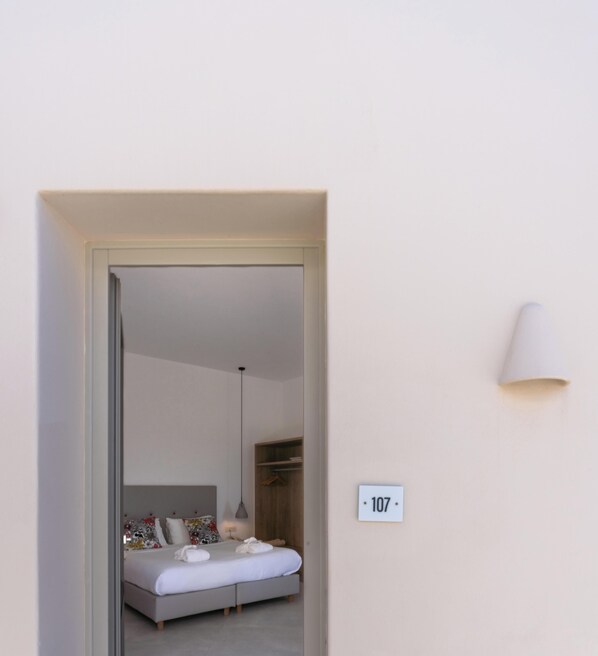 A bedroom with a bed, bedside table, lamp, and a wall-mounted number plate. - Hotiday Santorini Oia (Santorini)