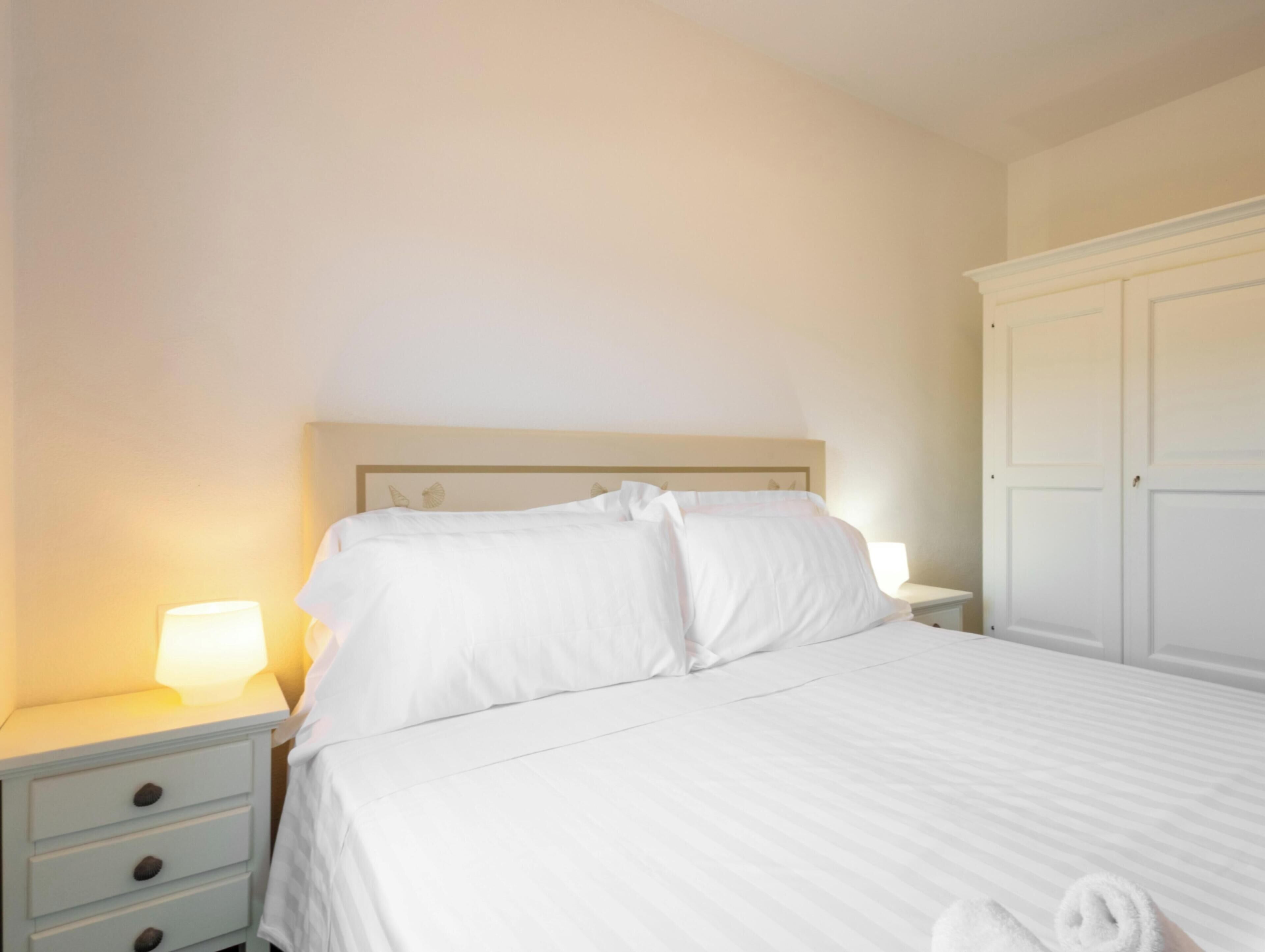 Un lit bien fait, avec du linge de lit blanc, une table de chevet avec une lampe et une armoire.
