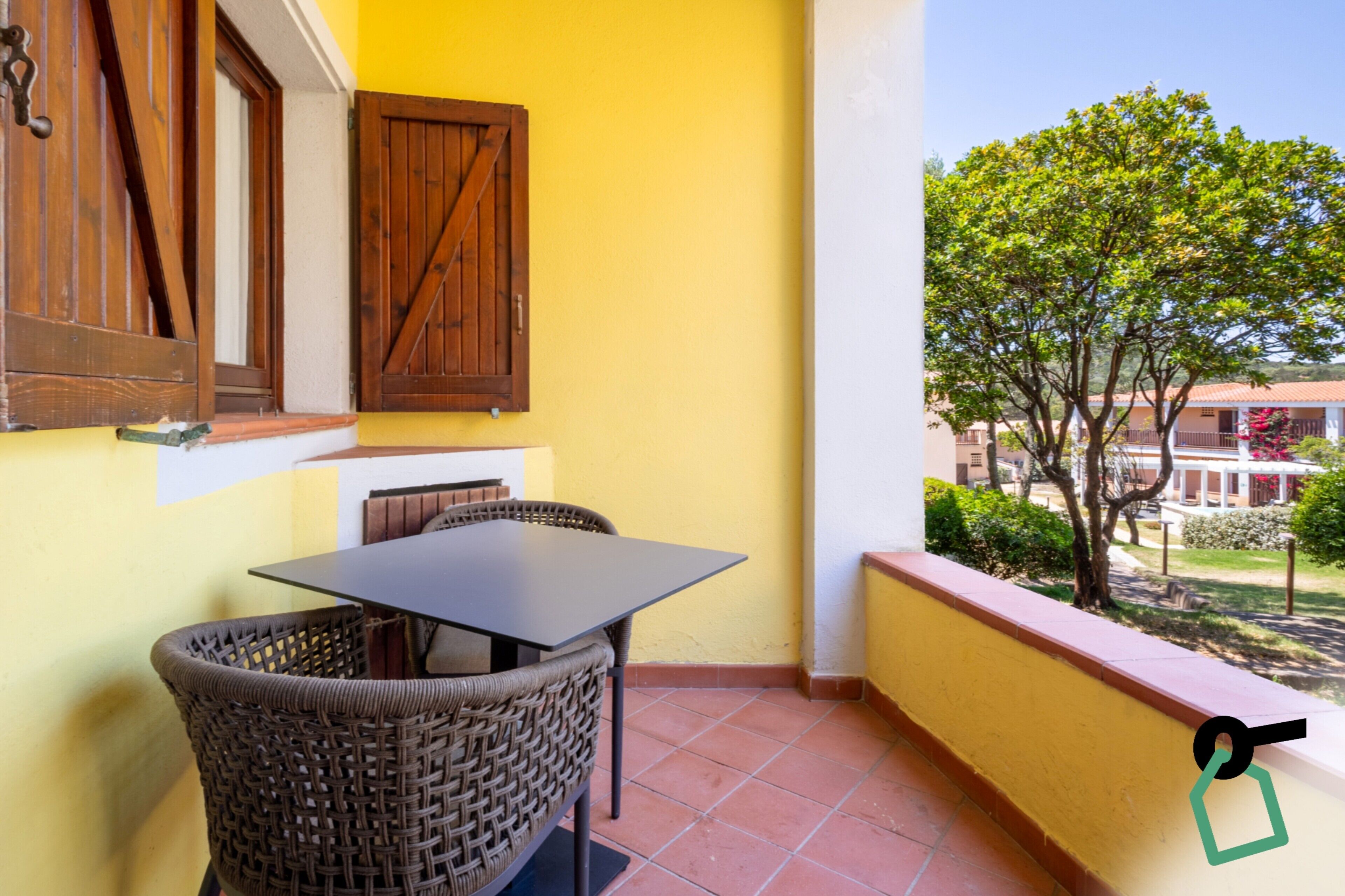 A balcony with a table and wicker chairs, a wooden door, and a view of a tree and buildings.