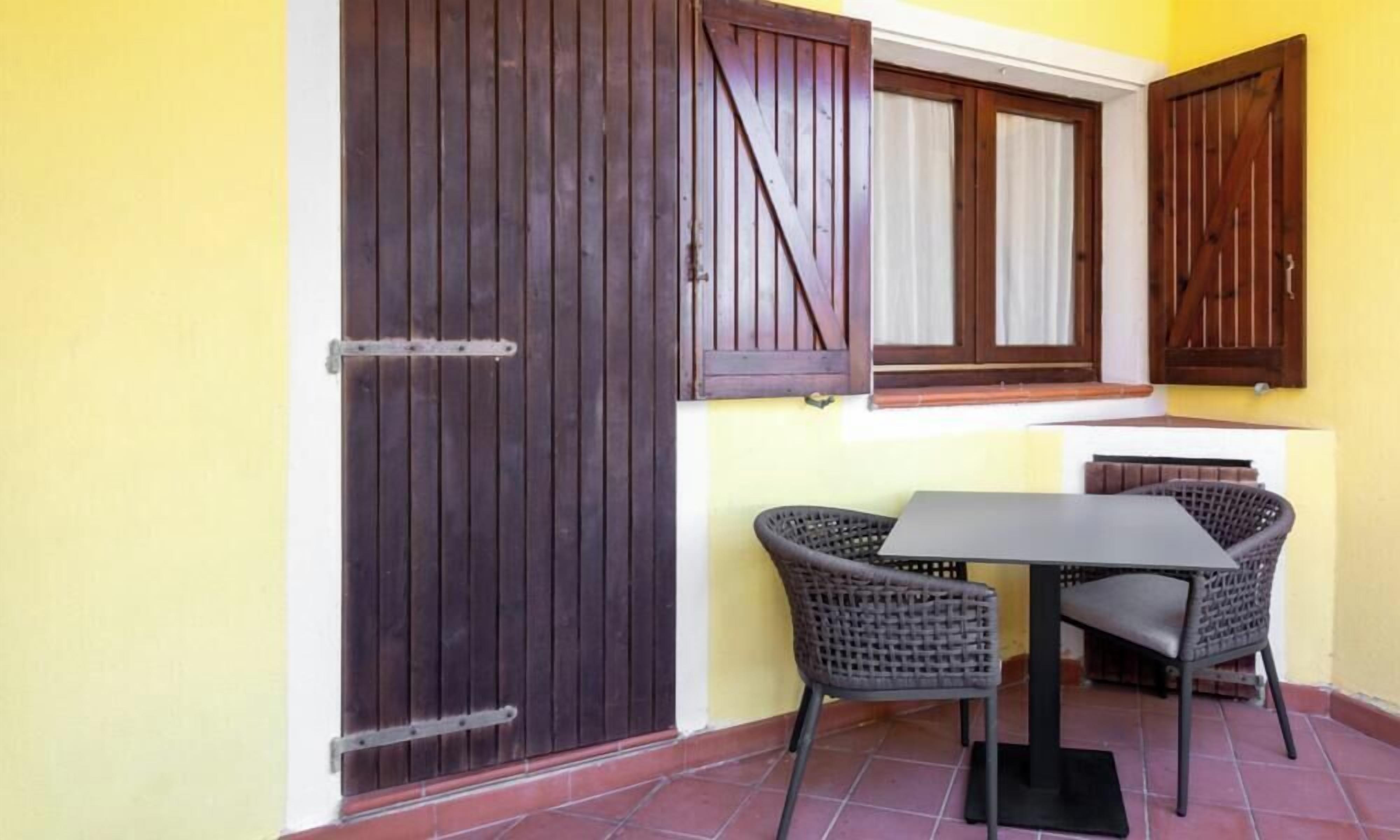 Une terrasse avec une table et deux chaises en osier, une porte en bois et une fenêtre avec des volets.
