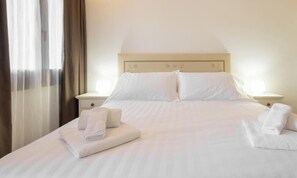 A neatly made bed with white linens and pillows, two bedside tables with lamps, and a window with curtains. - Hotiday Punta Sardegna (Costa Serena)