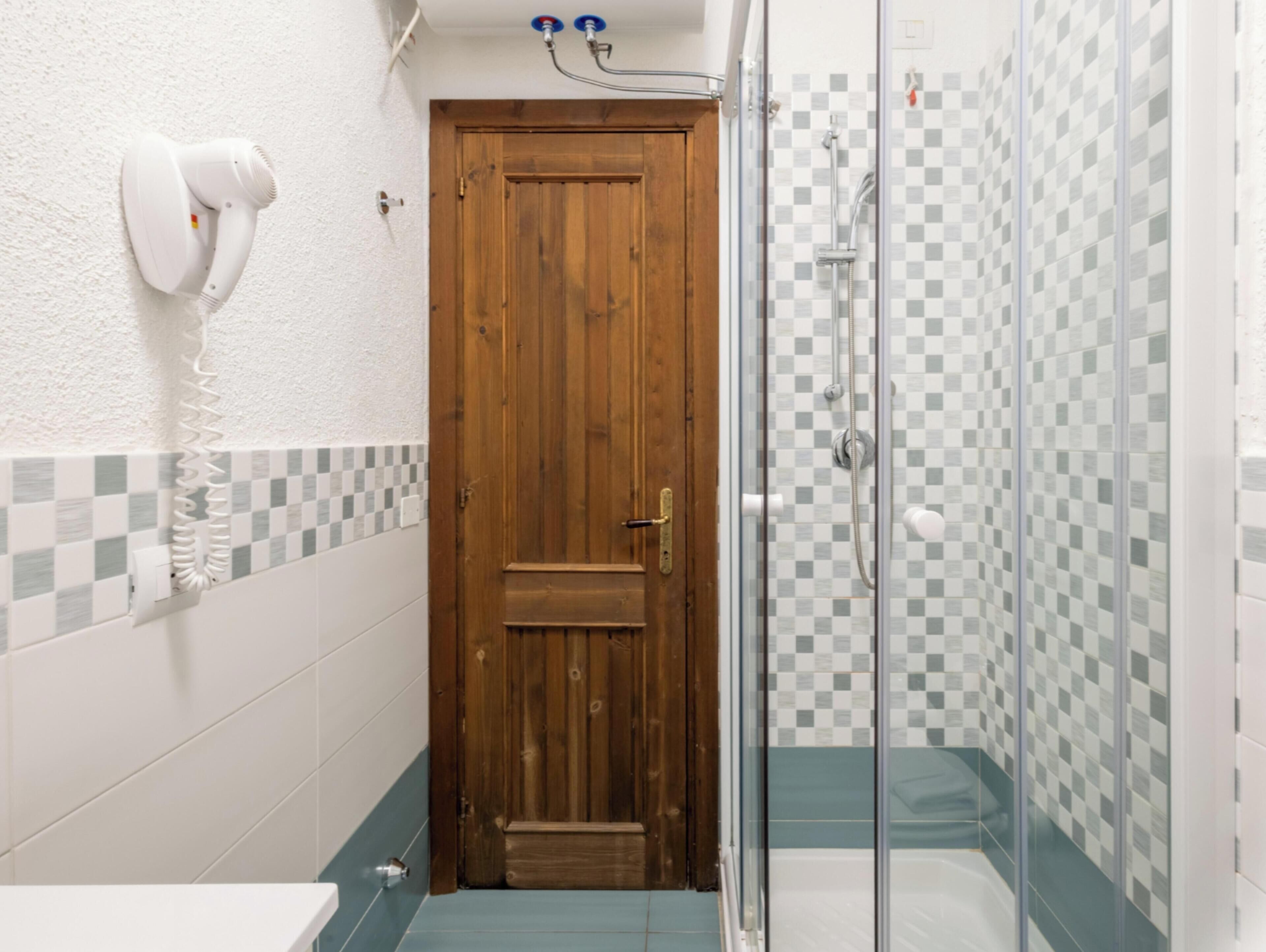 A bathroom with a wooden door, a shower with checkered tiles, and a white wall with a white towel dispenser.