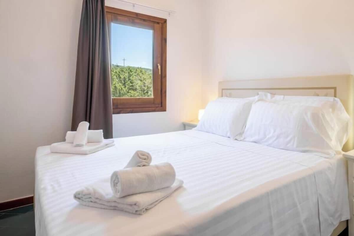 A neatly made bed with white linens, a window with a view of greenery, and a bedside table with a lamp.