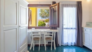 A room with a dining table and chairs, a window with curtains, and a view of the outside. - Hotiday Punta Sardegna (Costa Serena)