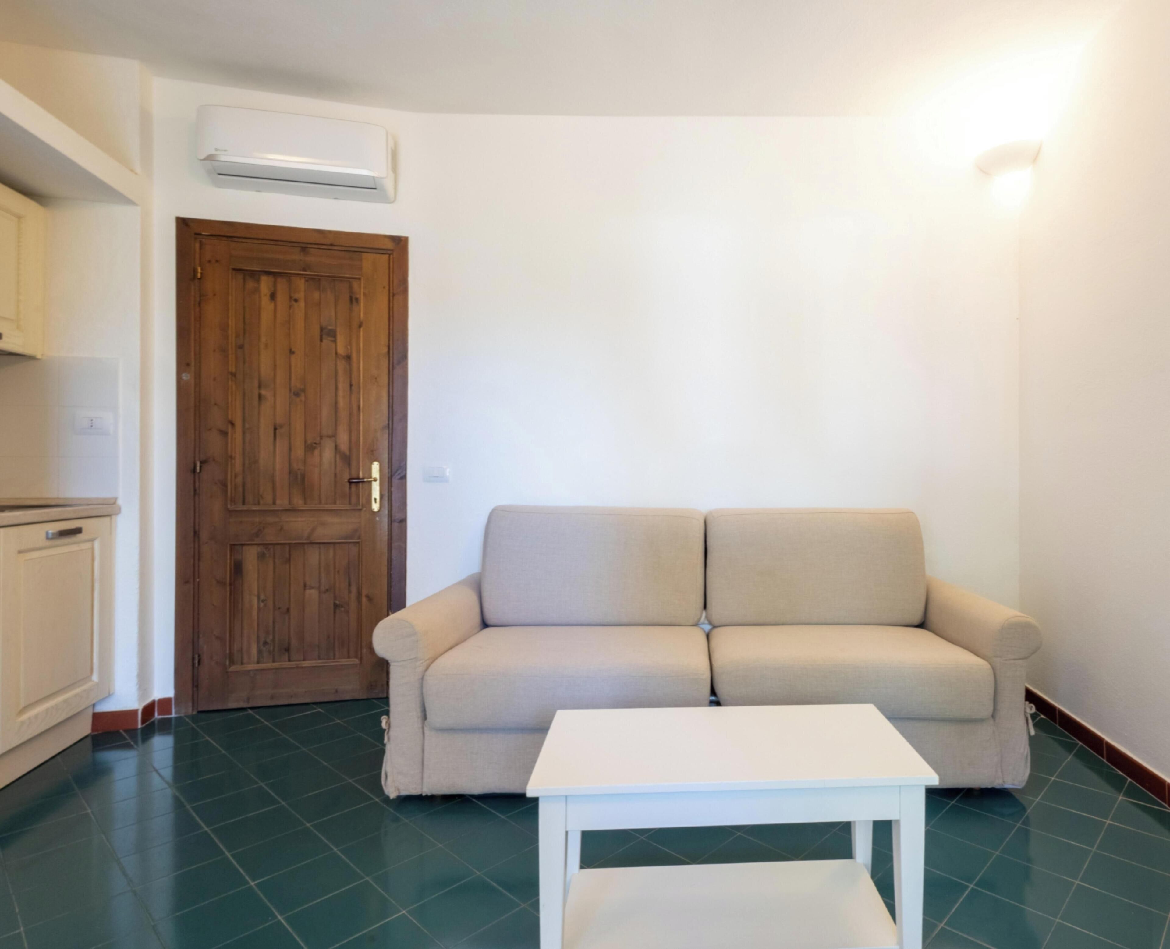 Une pièce avec un canapé beige, une table blanche, une porte en bois et un appareil de climatisation blanc.