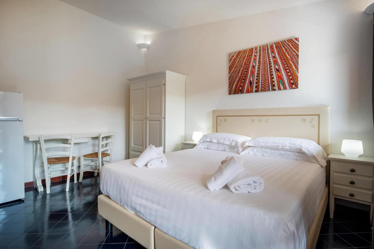 A hotel room with a bed, two white chairs, a desk, a wardrobe, and a wall-mounted artwork.