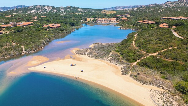An aerial view of a coastal area with a sandy beach, clear blue waters, and surrounding greenery. - Hotiday Punta Sardegna (Costa Serena)