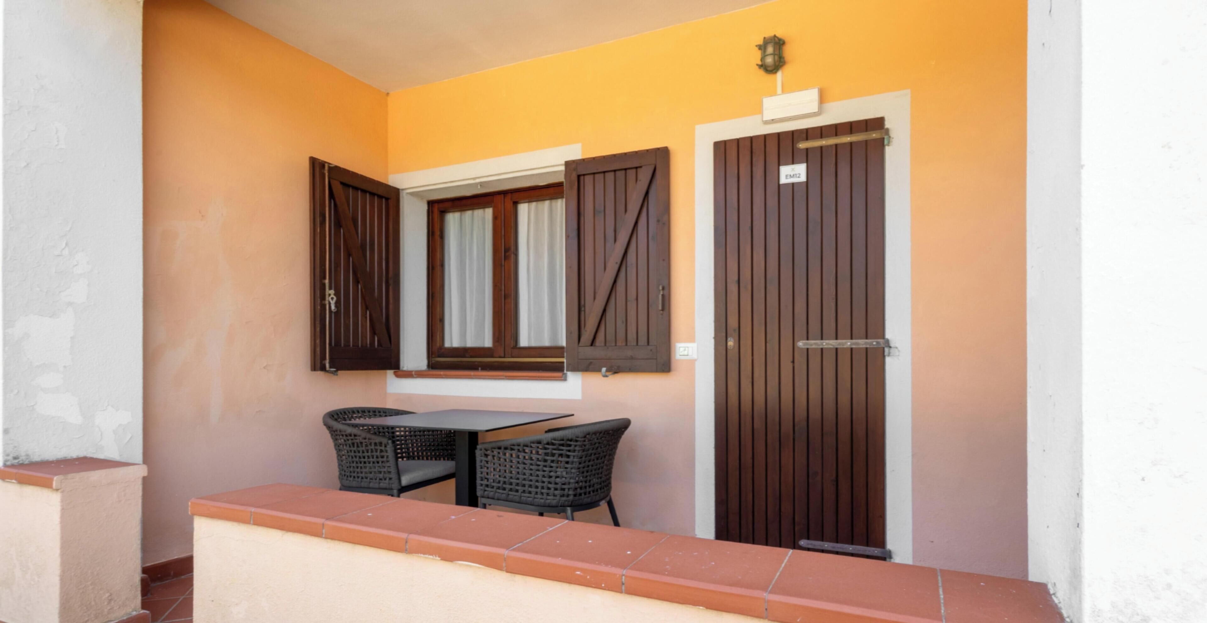 A balcony with two wicker chairs and a table, a wooden door with a sign, and a window with shutters.