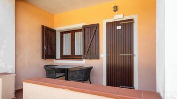 A balcony with two wicker chairs and a table, a wooden door with a sign, and a window with shutters.
