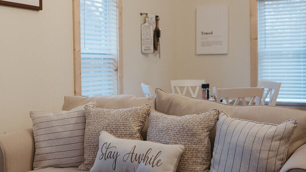 Un salon avec un canapé beige, des coussins à rayures et un coussin orné de la mention « Restez un peu ». Deux objets encadrés sont accrochés au mur, l’un avec du texte et l’autre avec une carte.