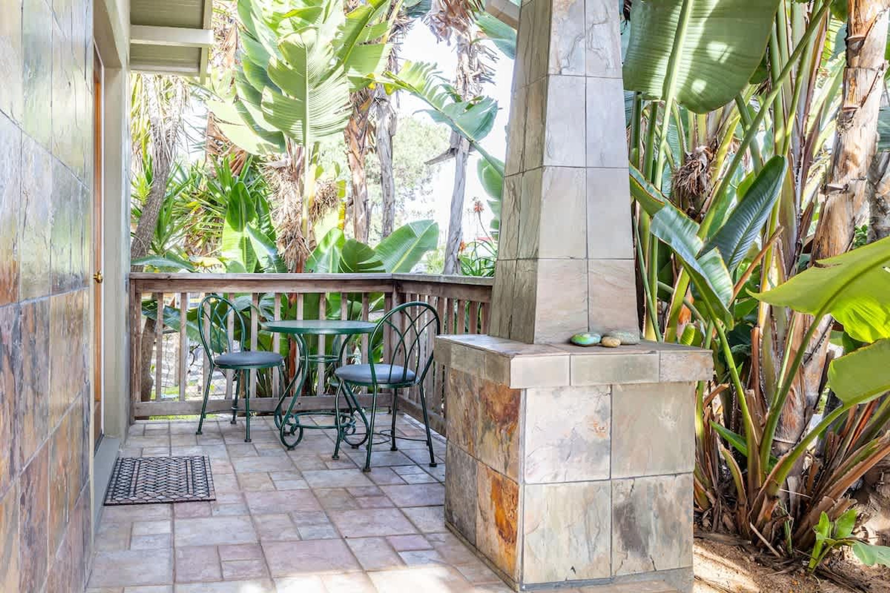 An outdoor patio with a tiled floor, two green metal chairs, a small round table, and a stone planter with a few rocks.