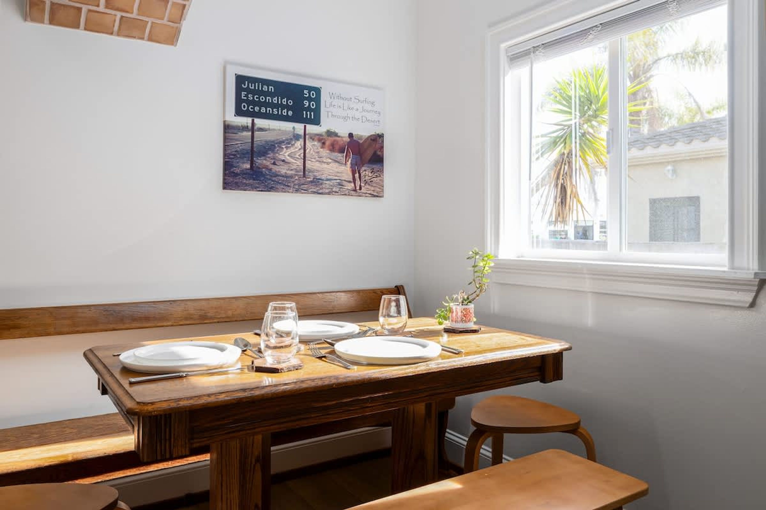 Un coin repas avec une table en bois dressée pour deux personnes, un banc et une fenêtre donnant sur des palmiers.