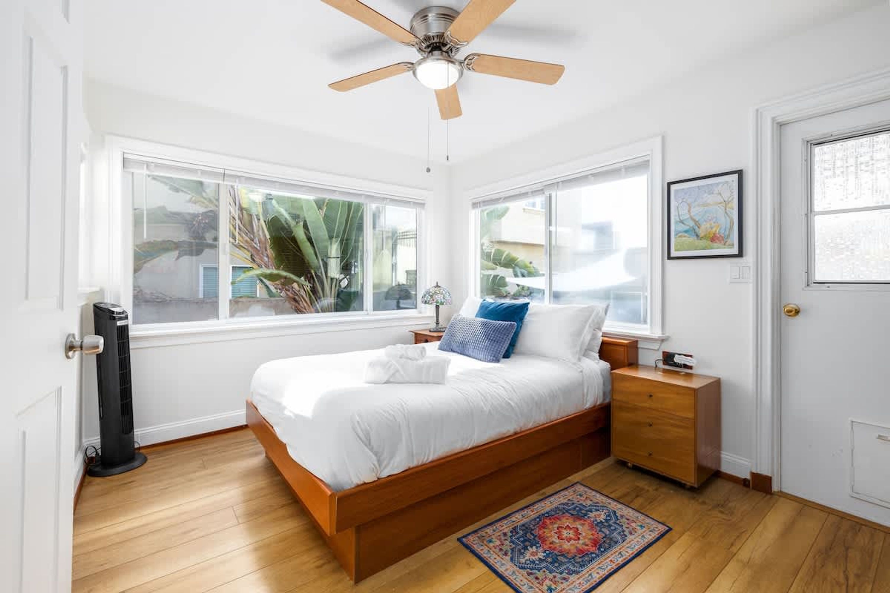 A bedroom with a bed, a ceiling fan, a nightstand, a dresser, and a window with a view of palm trees.