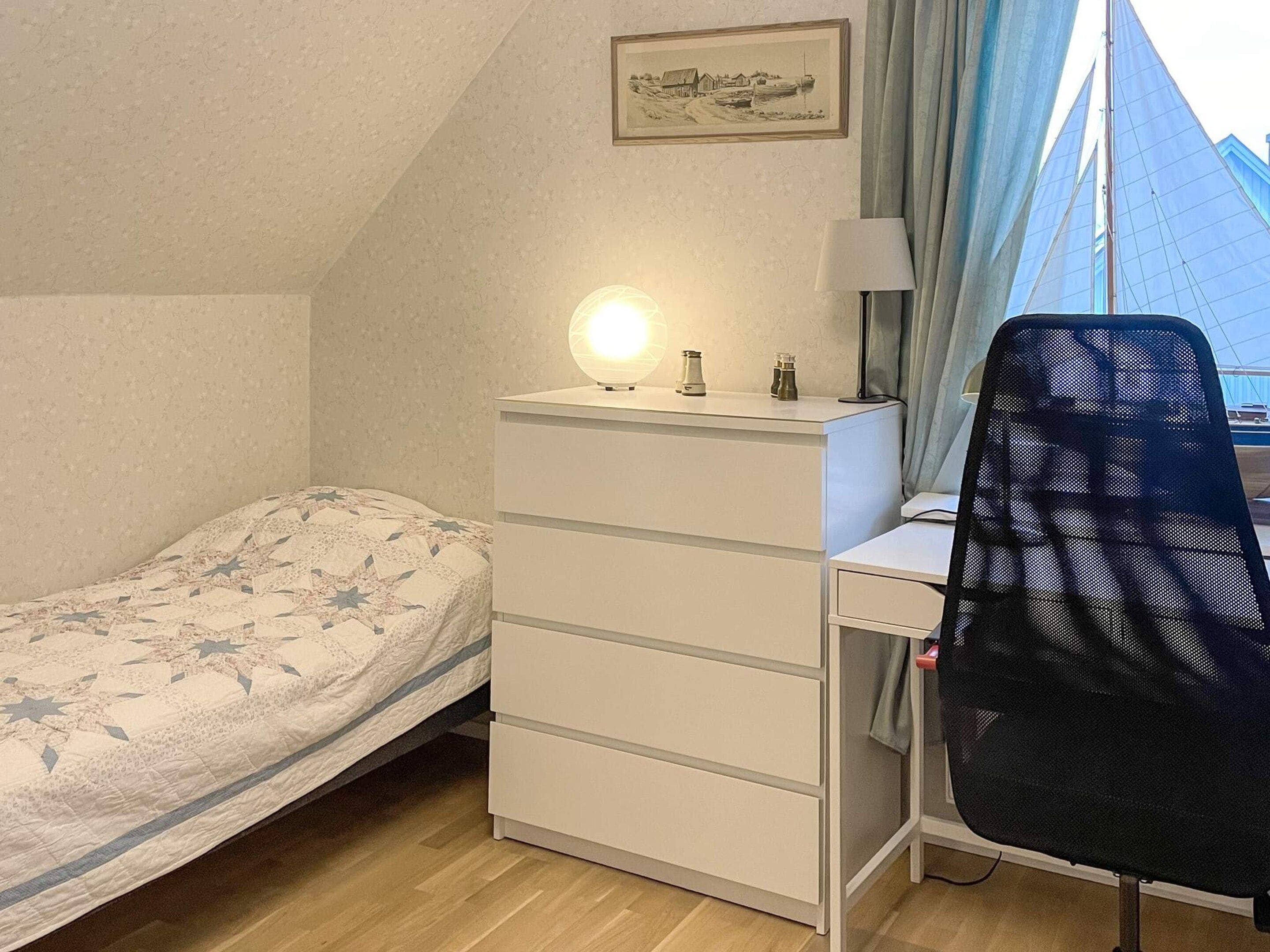 Ein Schlafzimmer mit einem Bett, einem Schreibtisch mit Stuhl, einem Schminktisch, einer Lampe und einem Fenster mit Vorhängen.
