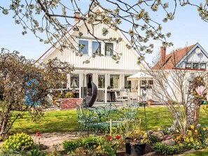 Una casa de dos pisos con un amplio jardín delantero, un comedor con mobiliario exterior y un jardín con diversas plantas y flores.