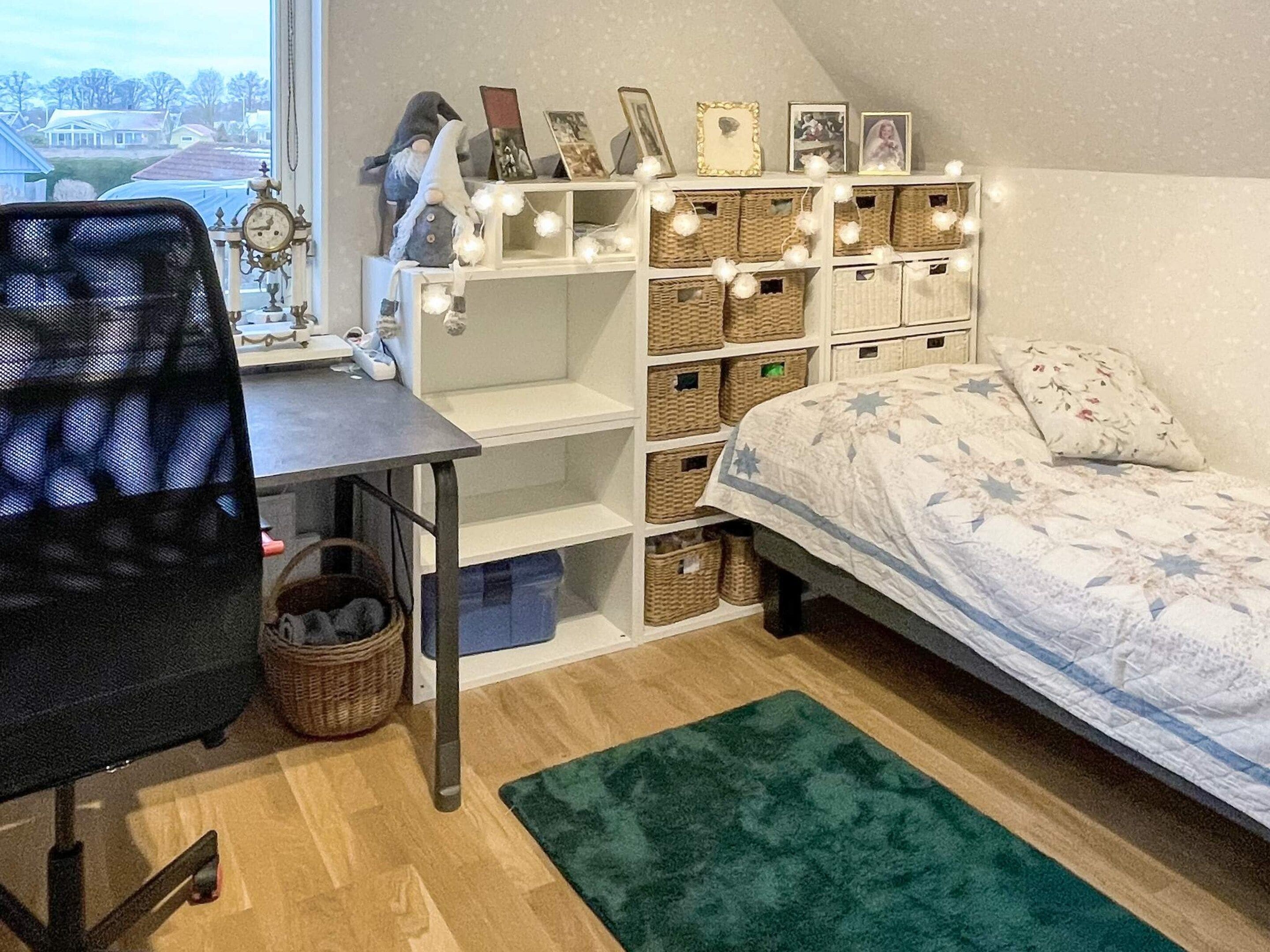 Et soveværelse med seng, skrivebord med stol, hylder med opbevaringskurve og et vindue med udsigt til huse.