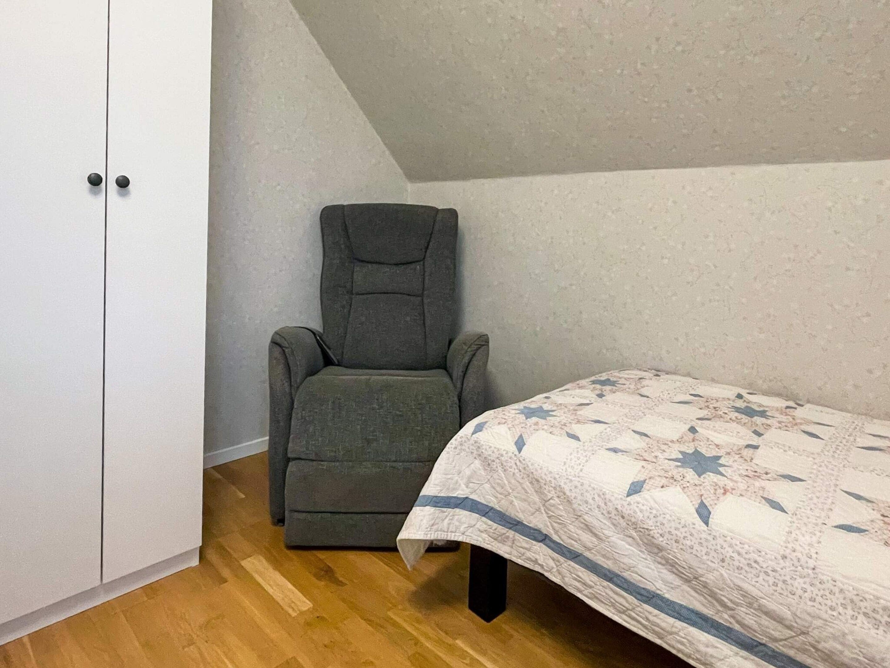 A bedroom with a single bed, a grey armchair, and a white wardrobe.