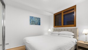 Un dormitorio con una cama, una mesita de noche, una lámpara, una ventana con persianas y un cuadro en la pared.