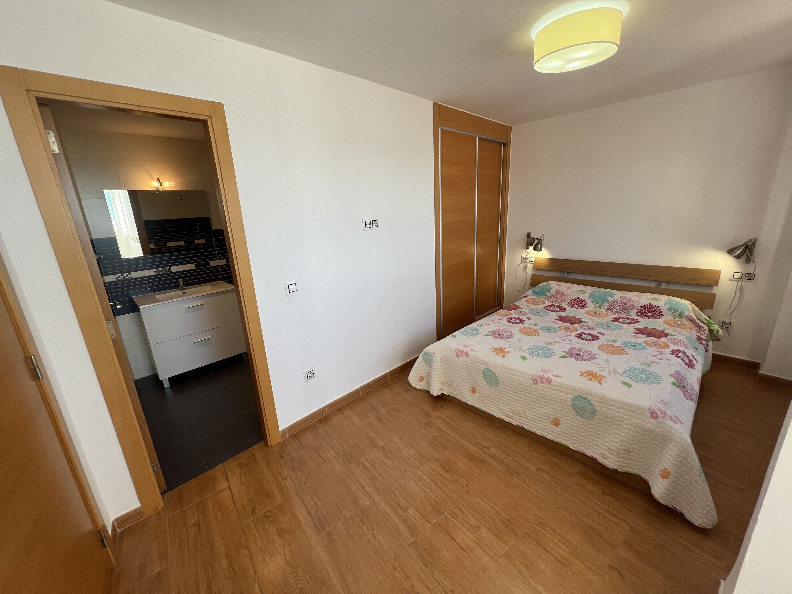 A bedroom with a wooden floor, a bed with a floral bedspread, a wooden wardrobe, and a bathroom with a tiled wall and a mirror.