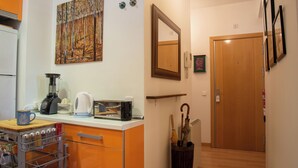 Una cucina compatta con macchina da caffè, tostapane e frullatore sul piano di lavoro. Uno scaffale in legno sopra il piano di lavoro ospita una foto incorniciata. Si vedono in secondo piano una porta in legno e uno specchio.