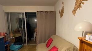 Una camera da letto con un letto ricoperto di beige, due cuscini rossi, un comodino in legno con una lampada, un tappeto decorato, una sedia blu e una porta a vetri scorrevoli che dà su un balcone.