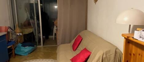 A bedroom with a bed covered in beige, two red pillows, a wooden nightstand with a lamp, a patterned rug, a blue chair, and a sliding glass door leading to a balcony.