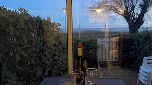 Un tavolo con una bottiglia e due bicchieri su un sottopiatti intrecciato, all'aperto con vista su un albero e una recinzione.