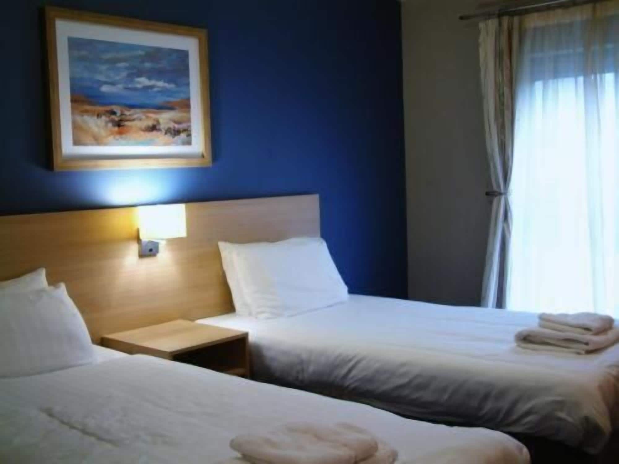 A hotel room with two beds, a nightstand, a painting on the wall, and a window with curtains.