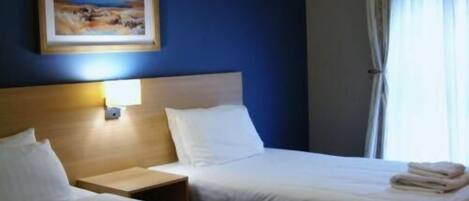 Una habitación de hotel con dos camas, una mesita de noche, un cuadro en la pared y una ventana con cortinas.