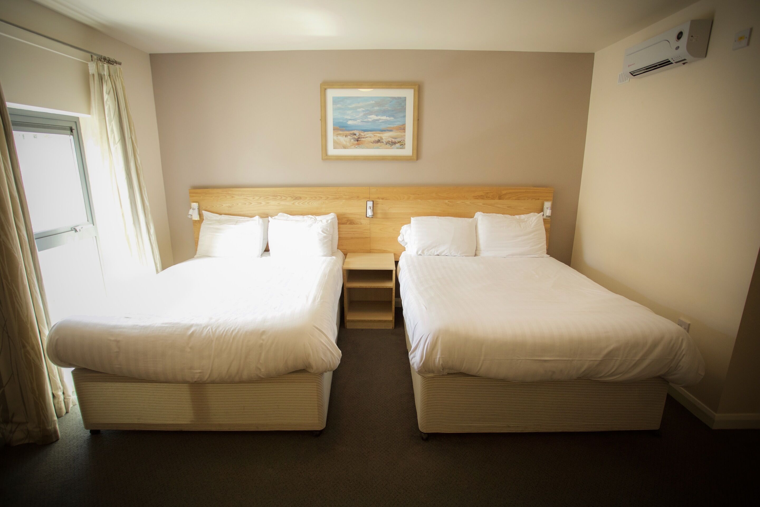 A hotel room with two beds, a wooden headboard, a framed picture on the wall, and an air conditioning unit.