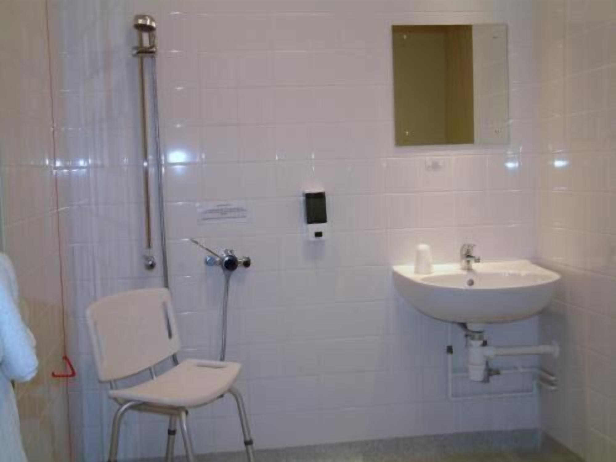 Un bagno con pareti rivestite di piastrelle bianche, un lavandino bianco, uno specchio e una doccia con soffione manuale.