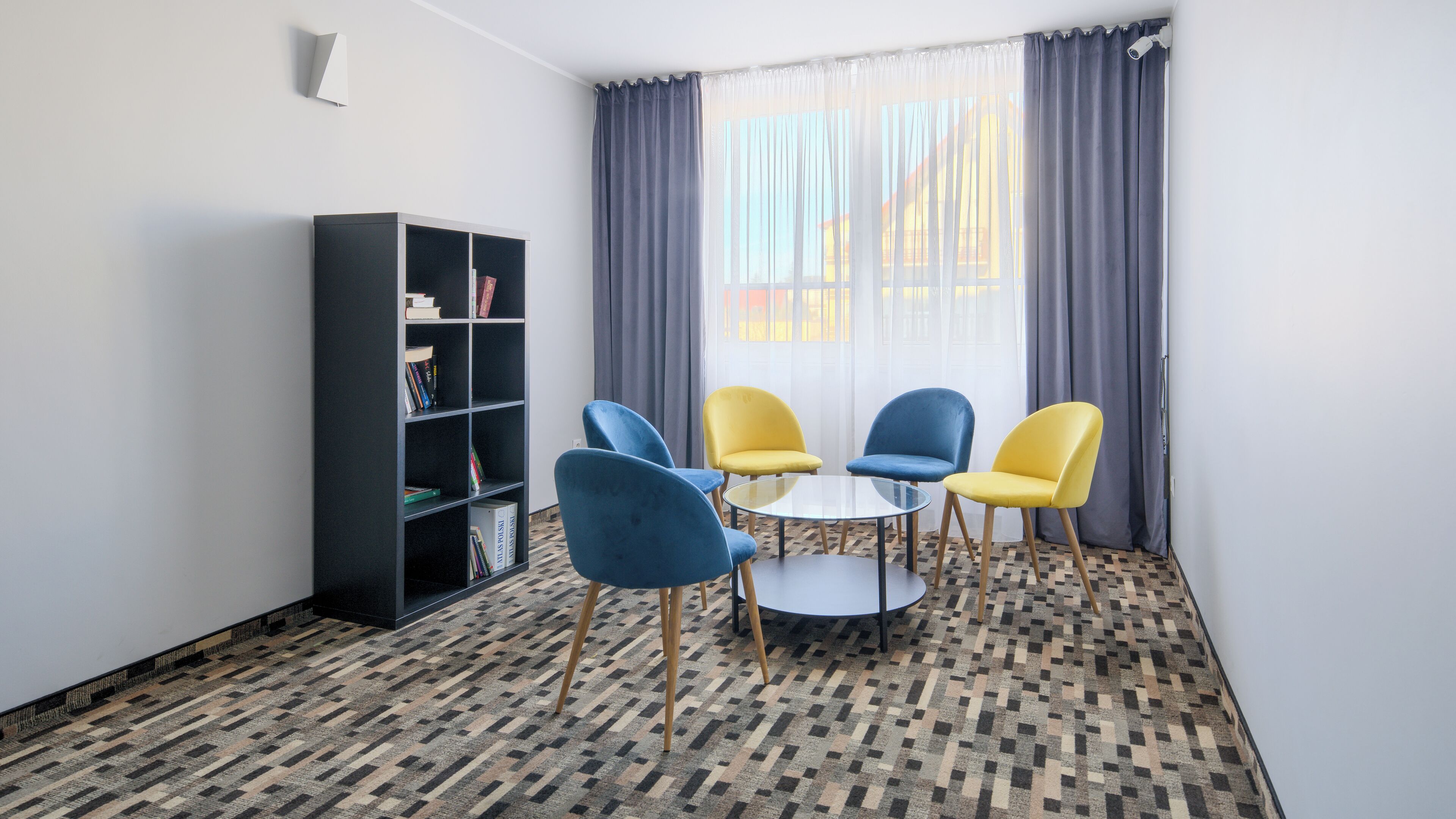 A room with a bookshelf, two blue chairs, one yellow chair, and a small round table.