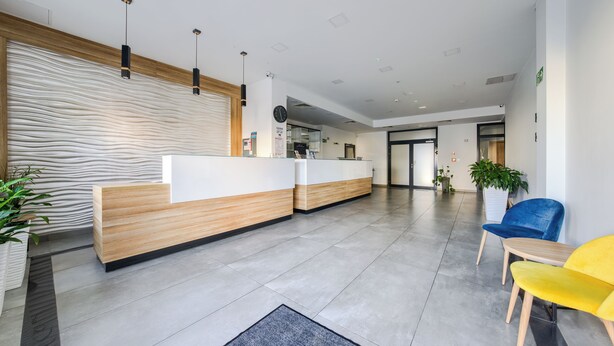 A modern reception area with a wooden reception desk, a clock, and a blue and yellow chair. - Apartamenty Sun & Snow Welle Resort (Grzybowo)