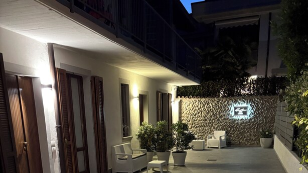 Un espace patio extérieur bien éclairé, doté d’un sol carrelé, d’un coin salon comprenant une chaise blanche et une petite table, ainsi que de plantes en pot.