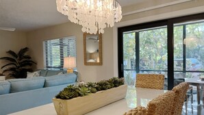 Uma sala de estar moderna com sofá, mesa de centro com plantas, cadeira de fibra natural, lustre e uma grande janela com vista para o verde.
