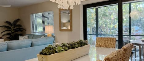 A modern living room with a sofa, a coffee table with plants, a woven chair, a chandelier, and a large window with a view of greenery.