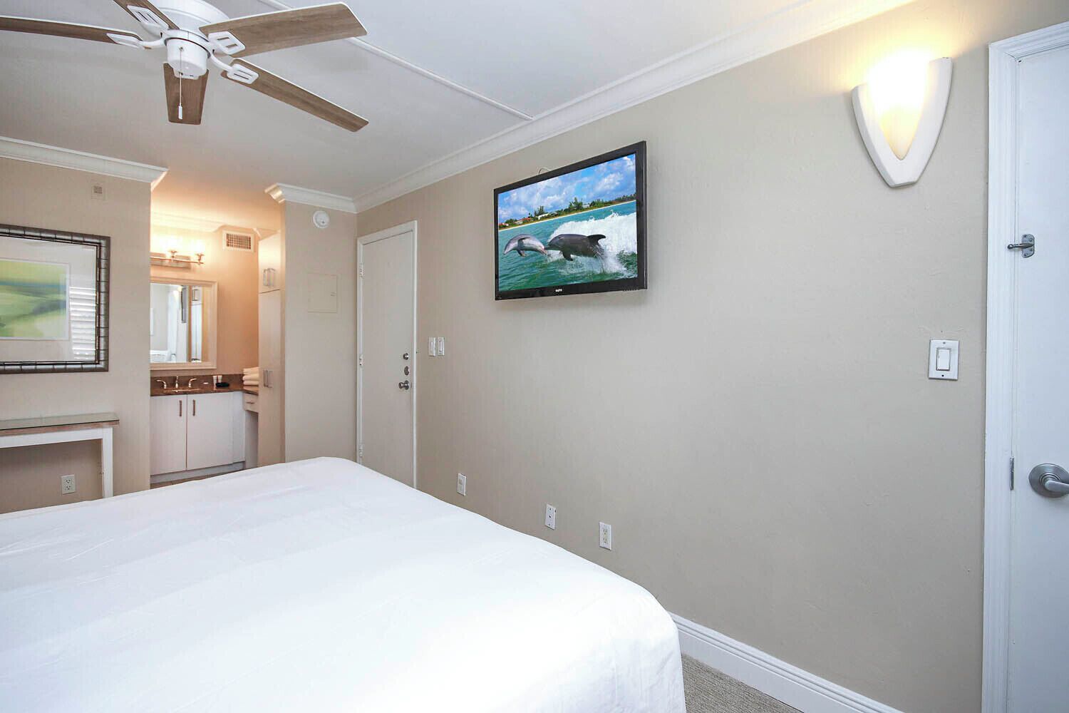 Habitación de hotel con cama, ventilador de techo, televisión montada en la pared y baño visible a través de una puerta abierta.