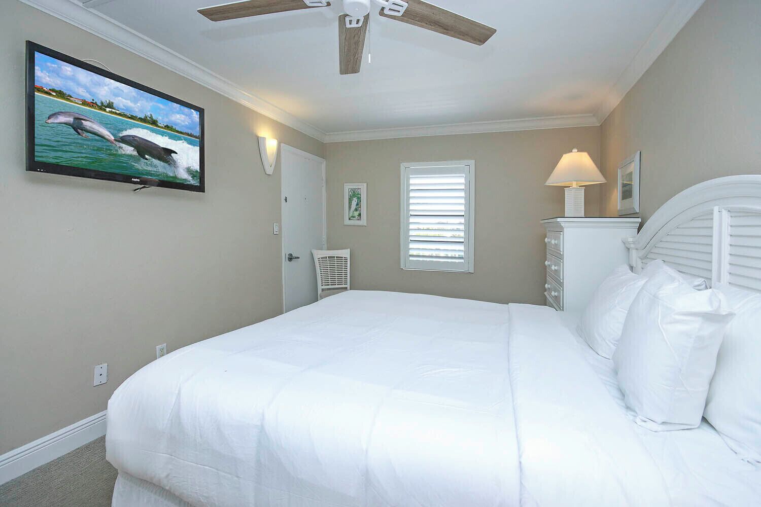 Un dormitorio con una cama, ventilador de techo, televisor en la pared, ventana con persianas y mesita de noche con lámpara.