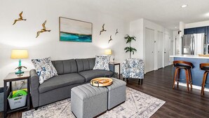 A modern living room with a grey sofa, matching ottoman, and a coffee table. There are two side tables with lamps and decorative items. A painting of a seascape hangs on the wall, and a plant is placed near the door. - Close to the Boardwalk, Jolly Roger Amusement Park and All the OC Fun! (Ocean City)
